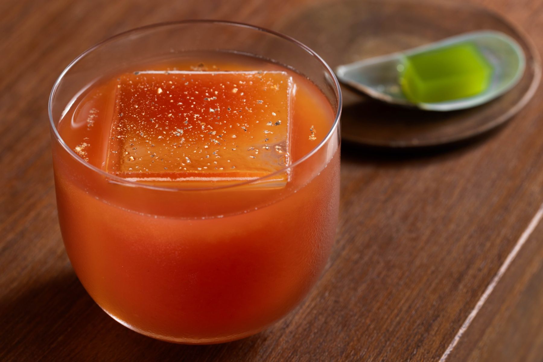 Red cocktail over a clear ice block with a green jelly on the side